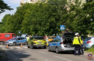 Na zdjęciu widoczne dwa radiowozy, pomiędzy nimi żółty samochód osobowy, który stoi na przejściu dla pieszych, widoczni policjanci w żółtych kamizelkach odblaskowych oraz osoby postronne.