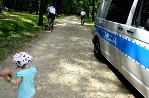 Policjanci na rowerach jadą w stronę lasu, widoczna dziewczynka oraz radiowóz.