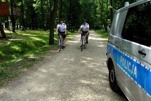 Dwaj umundurowani policjanci jadą na rowerach obok widoczny policyjny radiowóz.