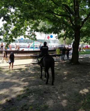 Policjant na koniu patroluje rejon jeziora Paprocany. Widoczne inne osoby oraz bawiące się dzieci.