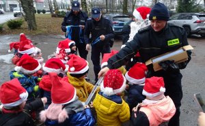 Funkcjonariusz Straży Miejskiej rozdaje odblaski dzieciom w czerwonych czapeczkach. Dalej widoczny mikołaj i policjanci.