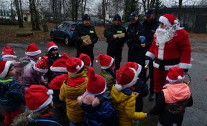 Mikołaj, strażnicy miejscy i policjanci a przed nimi grupka dzieci z czerwonymi czapeczkami.