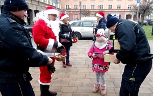 Jeden ze strażników miejskich daje dziewczynce opaskę odblaskową, widoczny Mikołaj i kobiety z czerwonymi czapeczkami.