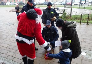 Mikołaj częstuje cukierkami dziecko, które stoi obok mamy, dalej policjanci i strażnicy miejscy.