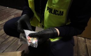 Funkcjonariusz w żółtej kamizelce w założonych rękawiczkach trzyma substancje psychoaktywne znajdujące się w woreczku.
