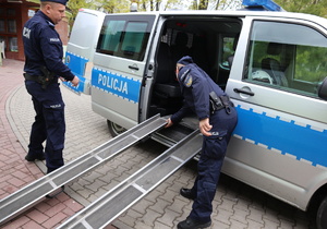 Dwóch umundurowanych policjantów montuje prowadnice do wjazdu wózków inwalidzkich, przy pomocy których podopieczni ośrodka wjadą do środka radiowozu.