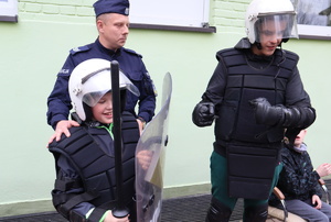 Podopieczni ośrodka ubrani w kamizelkę oraz hełm, jeden z nim trzyma tarczę, za nimi stoi umundurowany policjant.