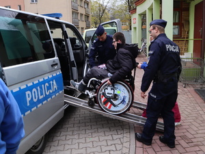 Policjanci pomagają osobie na wózku wjechać za pomocą prowadnic do policyjnego radiowozu.