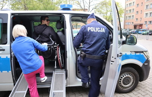 Chłopak na wózku znajduje się wewnątrz radiowozu za nim opiekunka, obok policjant.