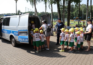 Dzieci wchodzą do policyjnego radiowozu, widoczne opiekunki oraz umundurowany policjant.