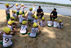 Dzieci na brzegu jeziora siedzą na piasku, przed nimi dwóch umundurowanych policjantów.