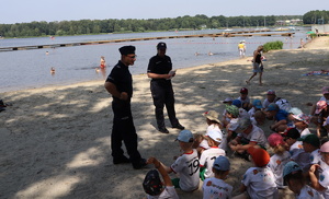 Policjanci stoją przed grupą dzieci, które siedzą na piasku przy brzegu jeziora.