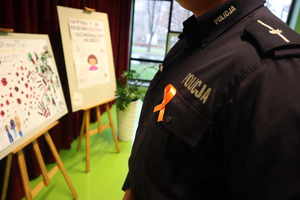 Widoczny mundur policjanta z napisem &quot;Policja&quot; oraz dopiętą pomarańczową wstążką, w tle widoczne prace plastyczne.