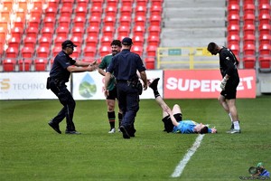 Na zdjęciu dwaj umundurowani policjanci na murawie boiska piłkarskiego. Obok nich trzech piłkarzy, jeden z nich leży na ziemi.