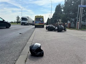Na zdjęciu kask motocyklowy oraz motocykl przewrócony na bok. W tle karetka oraz inne samochody jadące drogą.