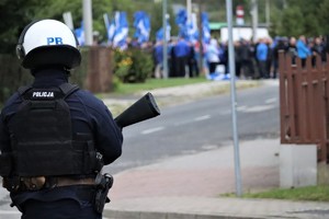 Na zdjęciu umundurowany policjant podczas zabezpieczenia meczu piłki nożnej. W tle grupa kibiców.