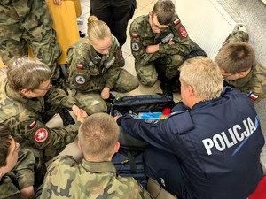 Na zdjęciu umundurowany policjant oraz uczniowie. Pomiędzy nimi torba medyczna.