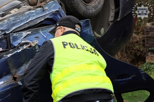 Na zdjęciu samochód uszkodzony w zdarzeniu drogowym oraz policjant w kamizelce odblaskowej z napisem Policja.