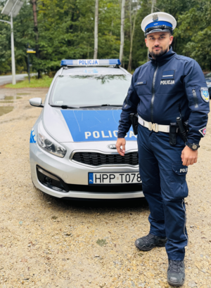 kolorowe zdjęcie umundurowanego policjanta Wydziału Ruchu Drogowego na tle oznakowanego radiowozu