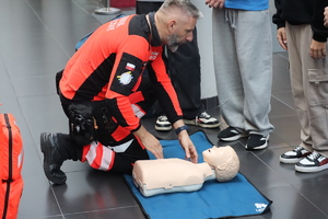 Kolorowe zdjęcie ratownika medycznego w towarzystwie młodzieży, który omawia zasady udzielania I pomocy.