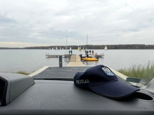 Kolorowe zdjęcie czapki policyjnej na podszybiu radiowozu, w tle widoczne jezioro.