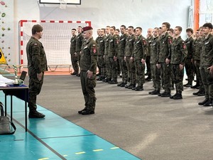 kolorowe zdjęcie umundurowanych drużyn składających się z uczniów klas mundurowych stojących w kolumnach na sali gimnastycznej. Na ich czele dowódca składający meldunek.