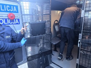 Kolorowe zdjęcie umundurowanych policjantów konwojujących zatrzymanych oznakowanym radiowozem.