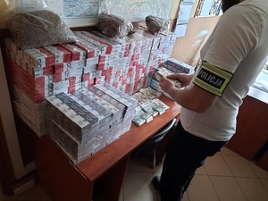Kolorowe zdjęcie stołu na którym znajdują się paczki papierosów oraz duża suma pieniędzy w nominałach 100 i 200 złotowych. Obok stoi nieumundurowany policjant, który przelicza pieniądze.