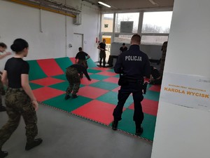 Kolorowe zdjęcie uczniów podczas ćwiczeń na matach na sali gmnastycznej. W tle widoczny umundurowany policjant stojący tyłem.