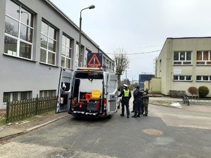 Kolorowe zdjęcie oznakowanego radiowozu policji w otoczeniu uczniów. Obok stoi umundurowany policjant Wydziału Ruchu Drogowego.