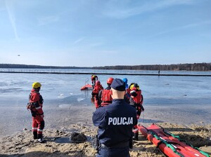 Kolorowe zdjęcie umundurowanego policjanta, w tle widać ratowników Wodnego Ochotniczego Pogotowia Ratowniczego stojących przy brzegu jeziora.