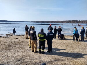 Kolorowe zdjęcie umundurowanych funkcjonariuszy Państwowej Straży Pożarnej, Policji oraz Wodnego Ochotniczego Pogotowia Ratunkowego. W tle widoczne jezioro.