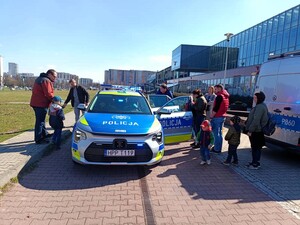 Dwa oznakowane radiowozy Policji, w pobliżu których znajdują się dzieci z opiekunami. W tle widać budynek Mediateki.