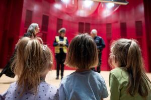 Troje dzieci uchwycone z tyłu, w tle widać scenę na której stoi Policjantka.