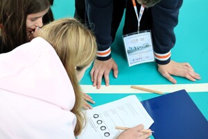 Uczniowie siedzący na podłodze sali gimnastycznej podczas rozwiązywania testu z wiedzy.