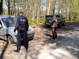 Umundurowany policjant oraz strażnik leśny wykonujący czynności w związku z zaparkowanym samochodem na terenach leśnych.