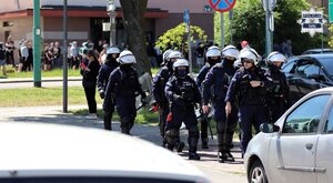 Policjanci w umundurowaniu oraz kaskach idący drużyną ulicą miasta. W tle widać kibiców.