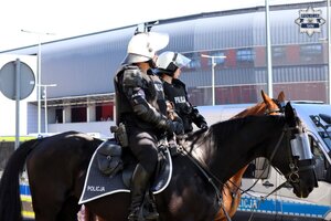Umundurowany policjant i policjantka na koniach. W tle widoczny oznakowany radiowóz oraz stadion miejski.