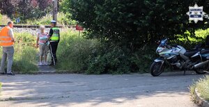 Pracownik Polskich Linii Kolejowych oraz umundurowany Policjant Wydziału Ruchu Drogowego rozmawiają z kobietą. W tle widoczne jest torowisko oraz oznakowany motocykl Policji.