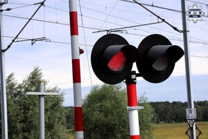Oznakowany przejazd kolejowy. Widoczne są podniesione rogatki oraz sygnalizacja świetlna nadająca sygnał koloru czerwonego.
