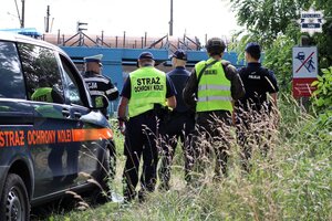 Umundurowani policjanci, pracownicy kolei oraz żołnierz Wojsk Obrony Terytorialnej stojących tyłem. W tle widoczne jest torowisko oraz samochód Straży Ochrony Kolei.