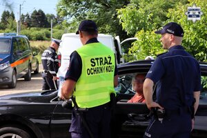 Umundurowani policjanci oraz pracownik Straży Ochrony Kolei rozmawiają z kierowcą samochodu.