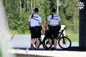Umundurowani policjanci patrolu rowerowego stojący w pobliżu brzegu jeziora. W tle widoczne rowery policyjne.