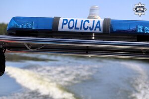 Listwa sygnałów świetlnych pojazdu uprzywilejowanego z napisem Policja znajdująca się na łodzi policyjnej. W tle widoczne fale na jeziorze, powstające za płynącą łodzią.