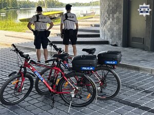Umundurowani policjanci patrolu rowerowego stojący w pobliżu brzegu jeziora. W tle widoczne rowery policyjne.