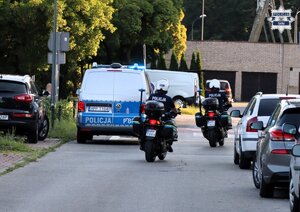 Umundurowani policjanci Wydziału Ruchu Drogowego jadący na motocyklach. W tle widoczny oznakowany radiowóz Policji.