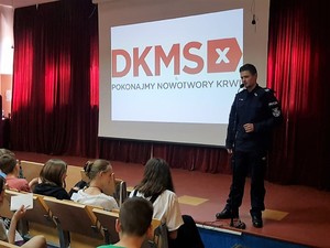 umundurowany policjant stoi na scenie, za sobą ma wyświetlone logo DKMS, przed nim, na widowni siedzi młodzież