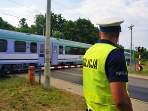 policjant w mundurze stoi przed przejazdem kolejowym, w tle widać opuszczone rogatki i przejeżdżający pociąg