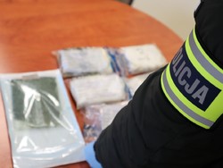 na stole leżą opakowania z narkotykami, przed nimi stoi policjant, który ma na ramieniu opaskę z napisem Policja