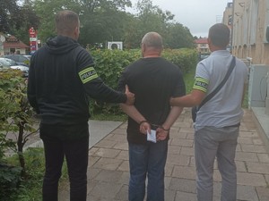 Dwaj nieumundurowani policjanci trzymają zatrzymanego mężczyznę, stoją tyłem do zdjęcia